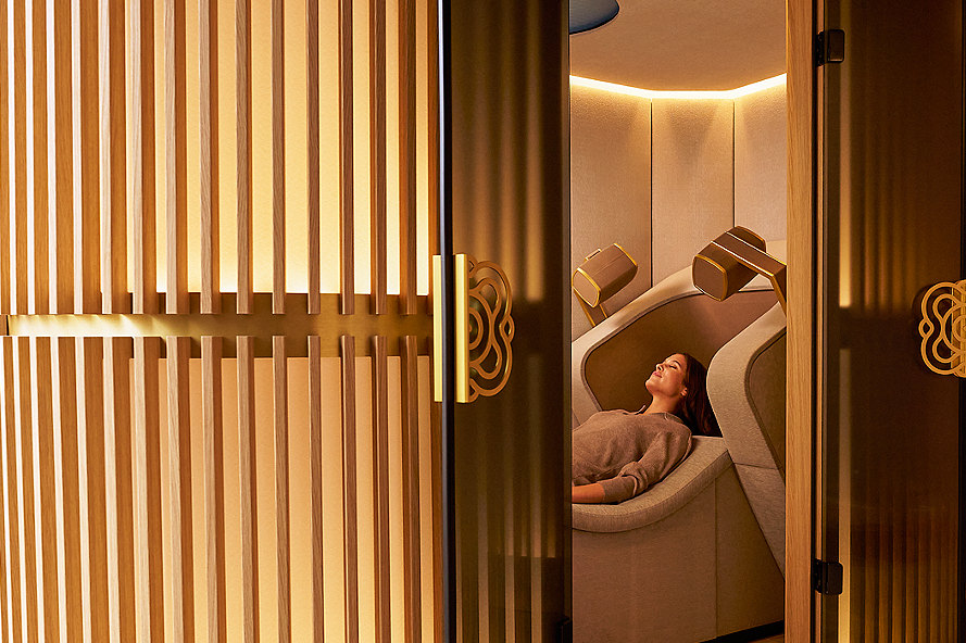 A Brain massage cabin with warm-lit wooden slats, doors open to a woman reclined in a zero-gravity chair