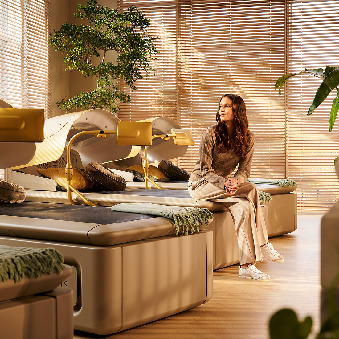 A woman sitting on the edge of a hydro massage bed in a sunlit Mind Oasis room, blinds and greenery behind her