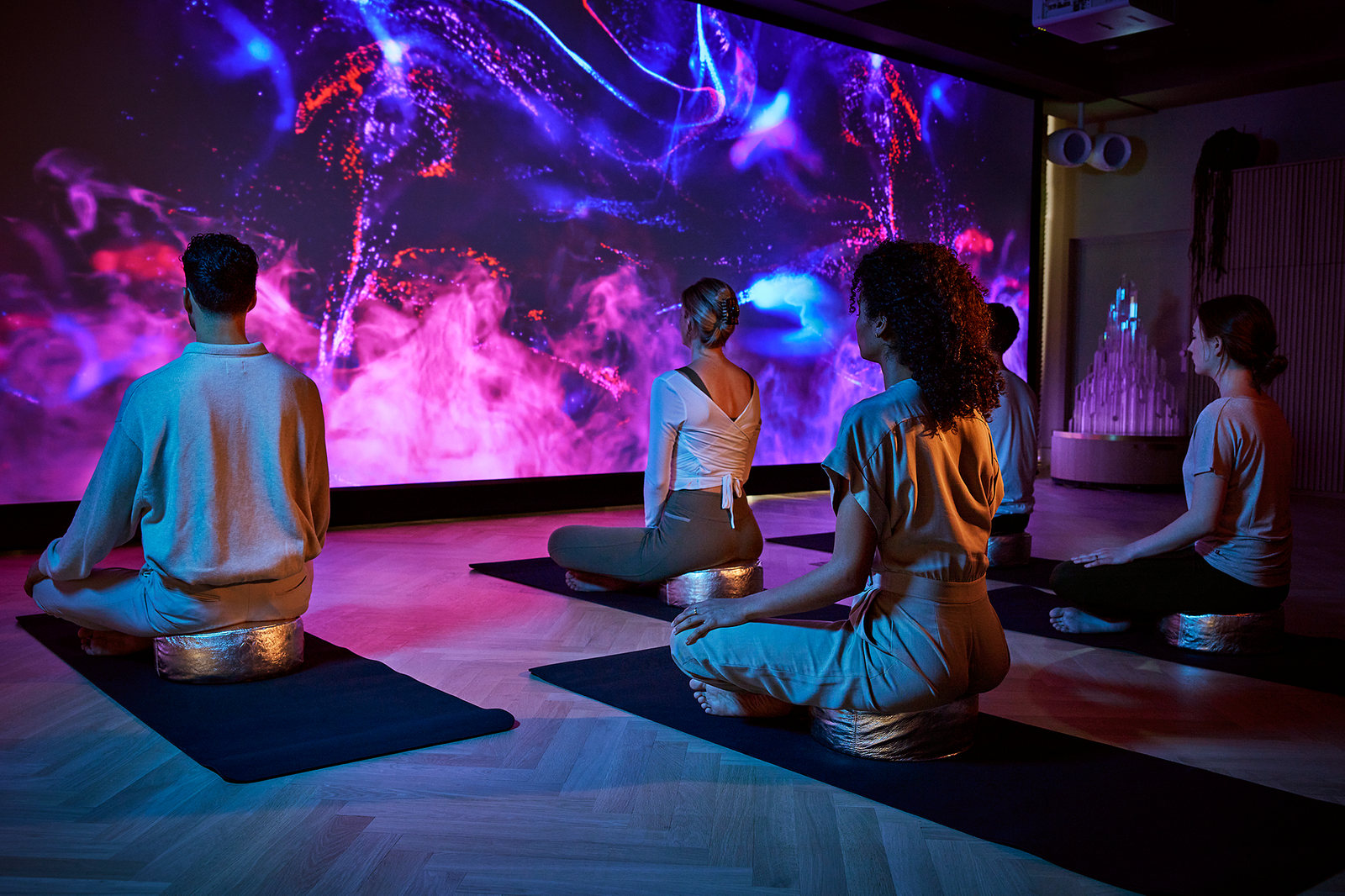 Four people meditating on mats in the House of Rituals mind spa, a wall-sized projection of pink and blue light in front of them