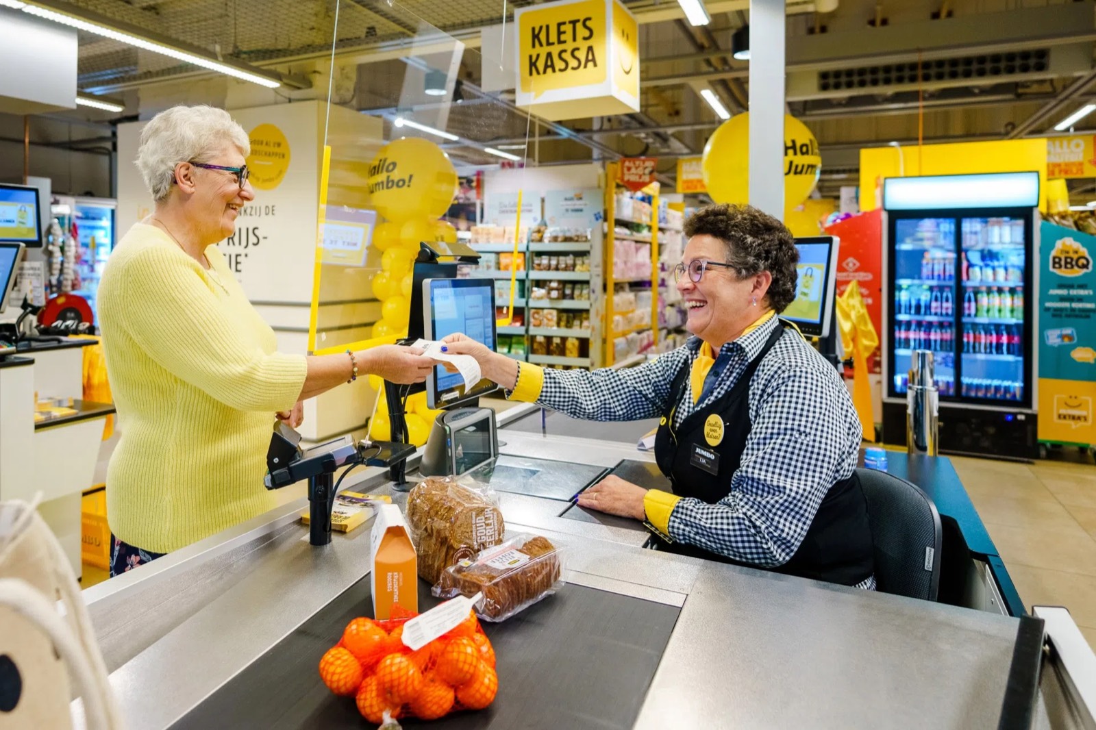 A Jumbo cashier and an older customer smiling at each other as they hand over a receipt at a checkout marked Kletskassa