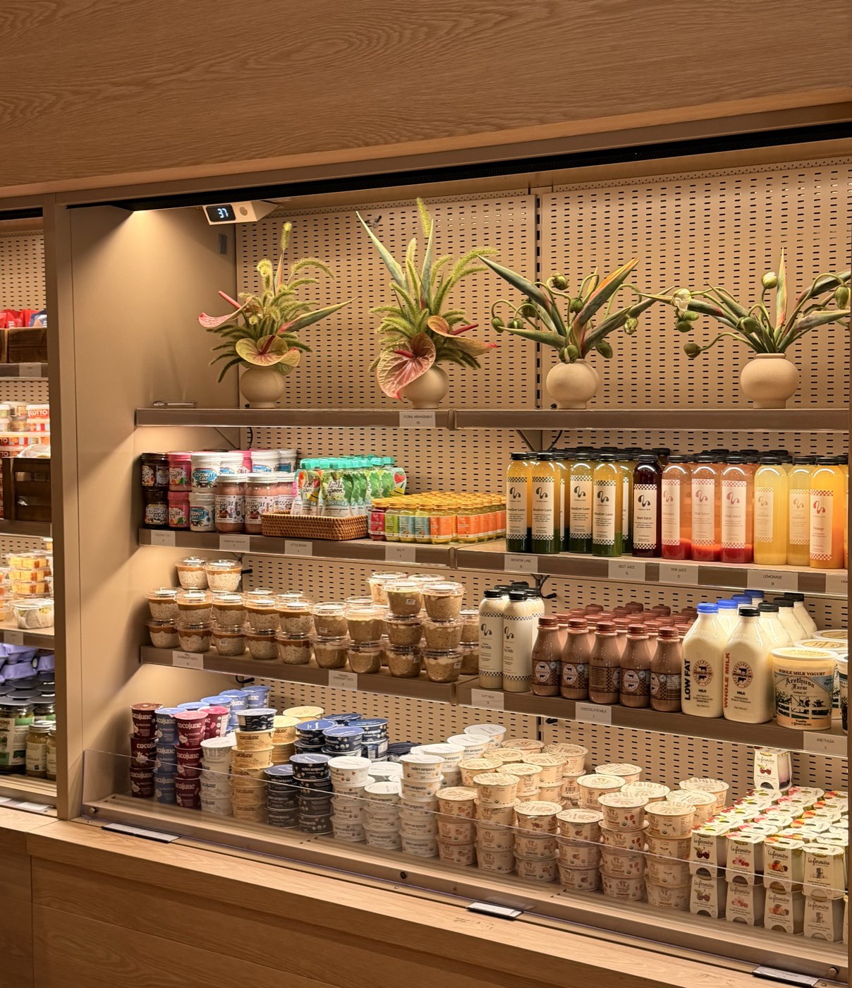 A chilled wall at Meadow Tribeca: pale oak and pegboard shelving lined with cold-pressed juices, yogurts and milk, with potted flowering plants on the top shelf