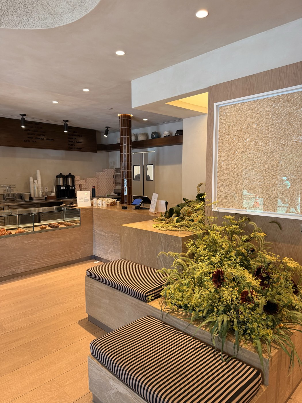 The front of Meadow Tribeca: a plaster coffee counter and pastry case beside travertine benches with striped cushions and an abundant arrangement of yellow blooms