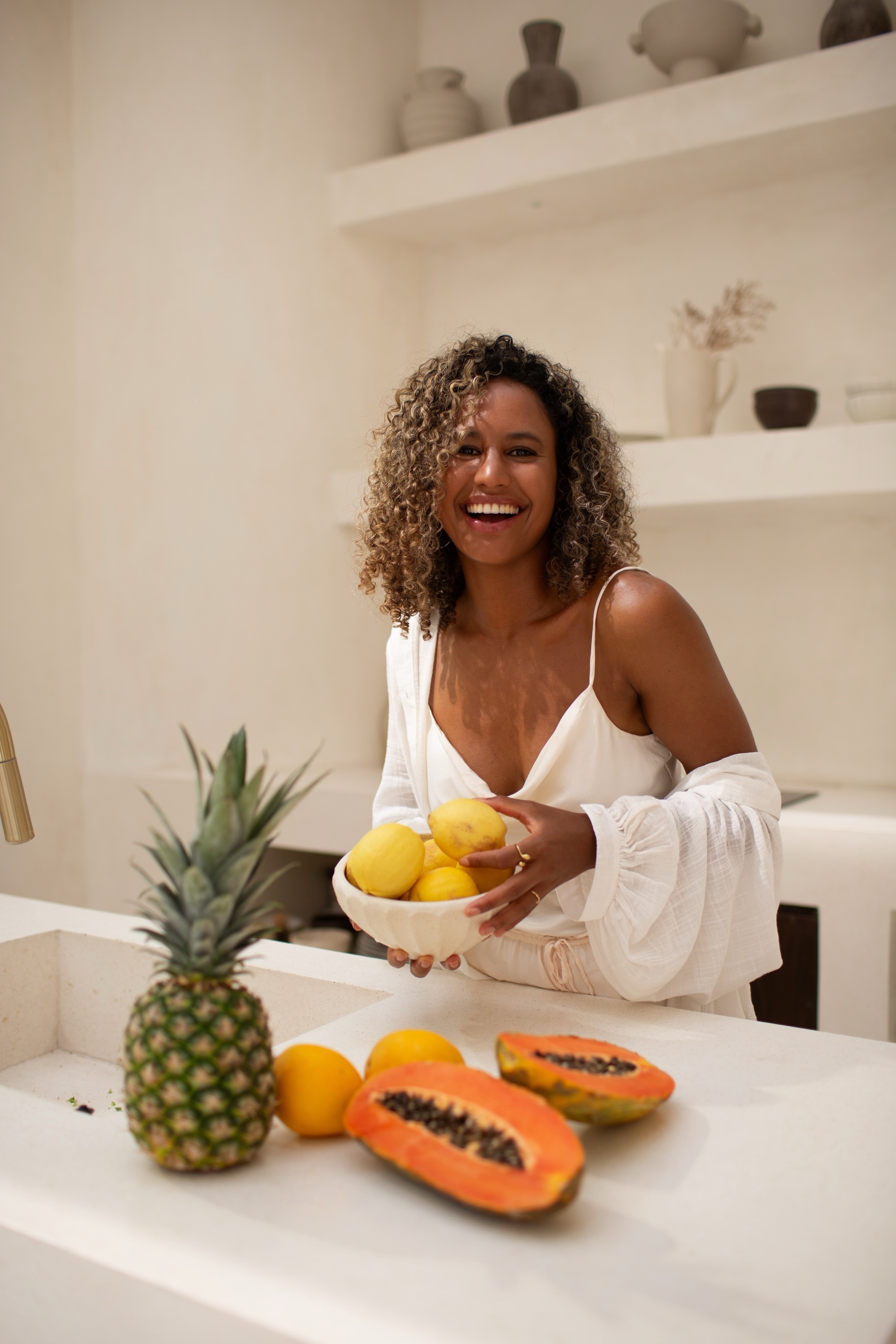 Couro in a bright kitchen holding a bowl of lemons, fresh fruit on the counter