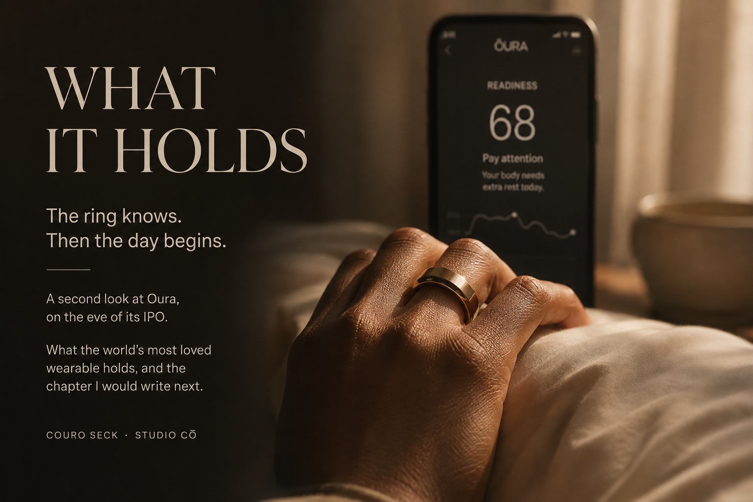 What It Holds — a hand wearing a gold Ōura ring resting on a bed beside a phone showing a readiness score of 68